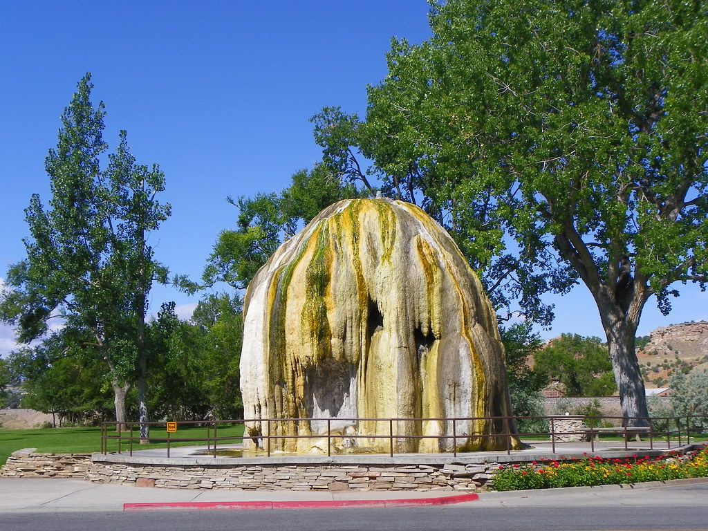 A large dome-shaped mineral hot spring formation surrounded by green trees in a park setting under a clear blue sky.