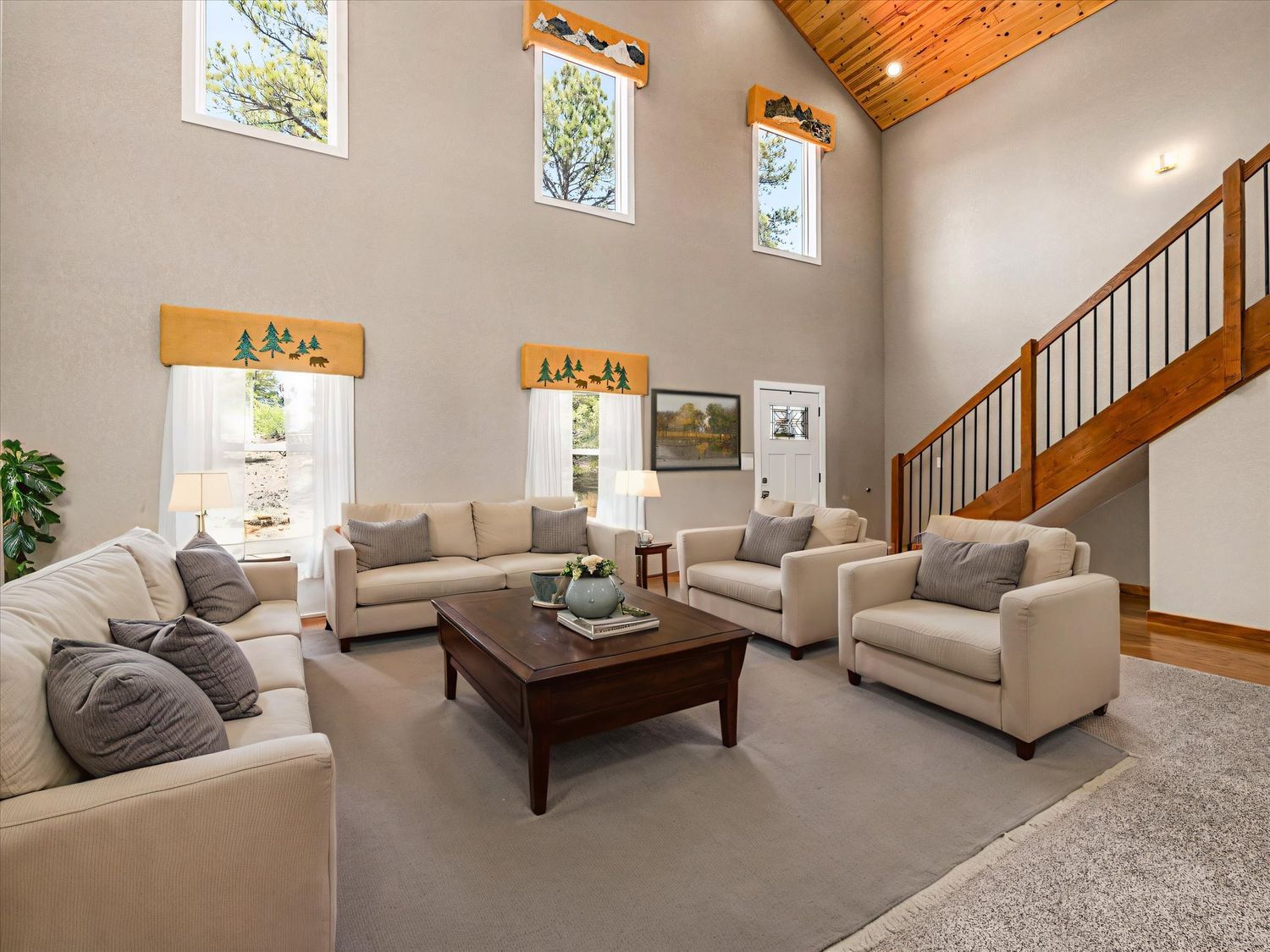 Spacious living room with vaulted wood ceiling, large windows, neutral sofas, and open staircase.