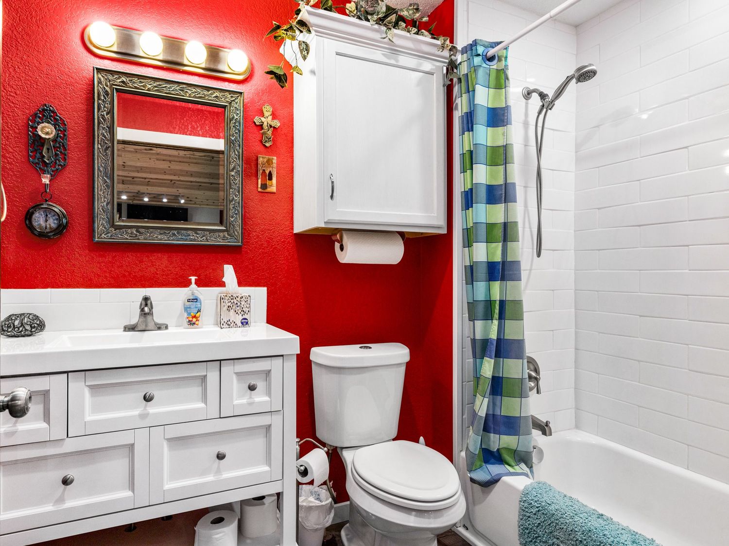 Bright red bathroom with white vanity, mirror with lights, toilet, and tiled shower with colorful curtain.