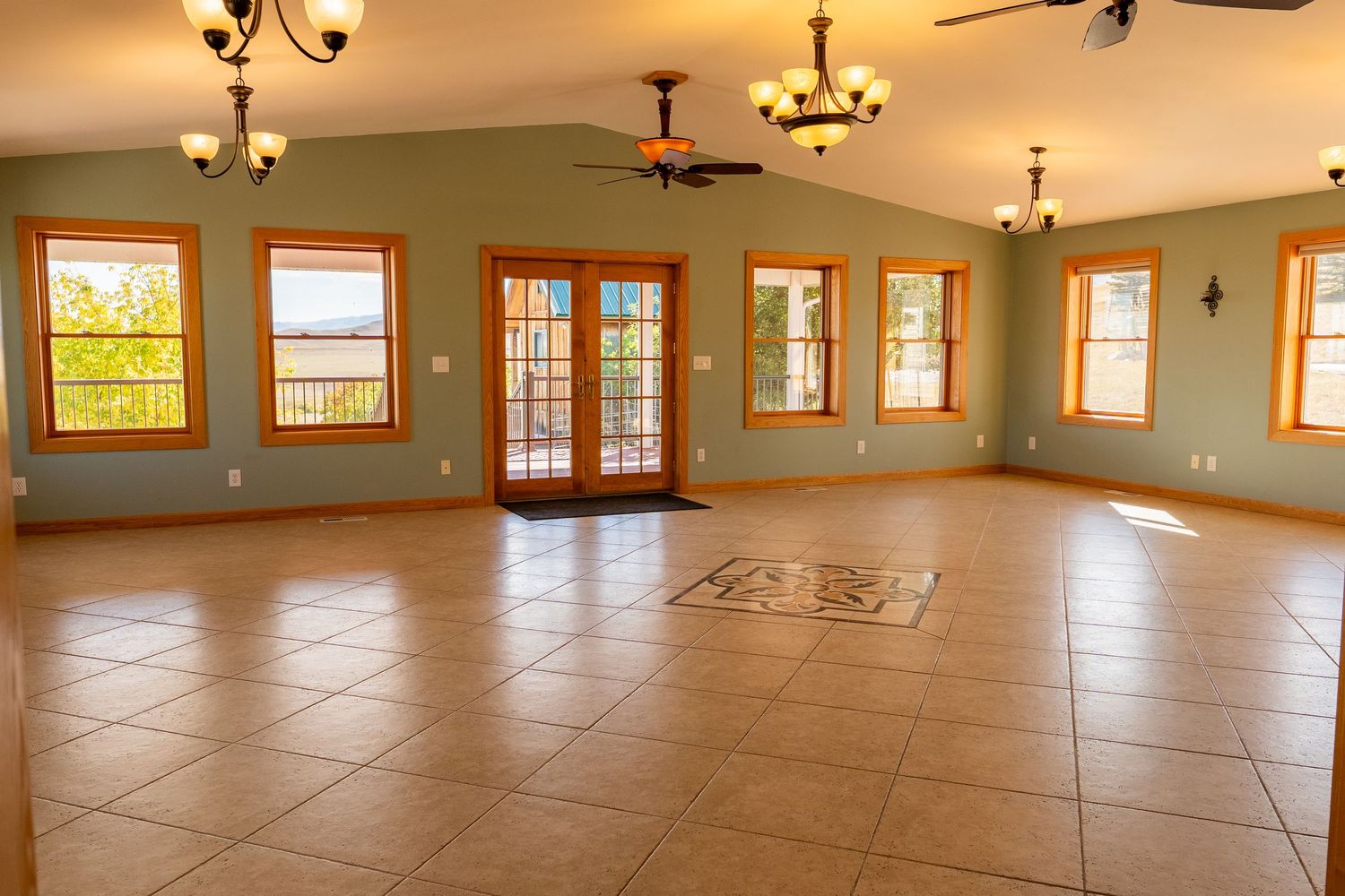 Spacious open living room with tile flooring, green walls, wood-trimmed windows, French doors, and ceiling fans.