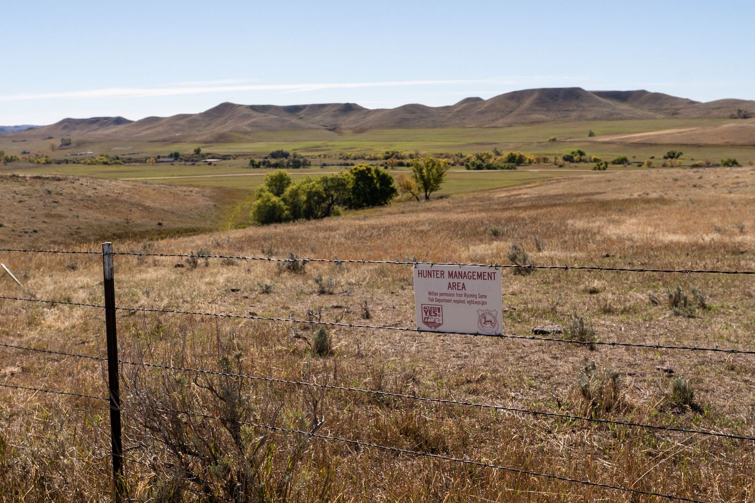 Vast open rangeland with a barbed wire fence and posted sign, rolling hills and green trees in the background under a clear sky.