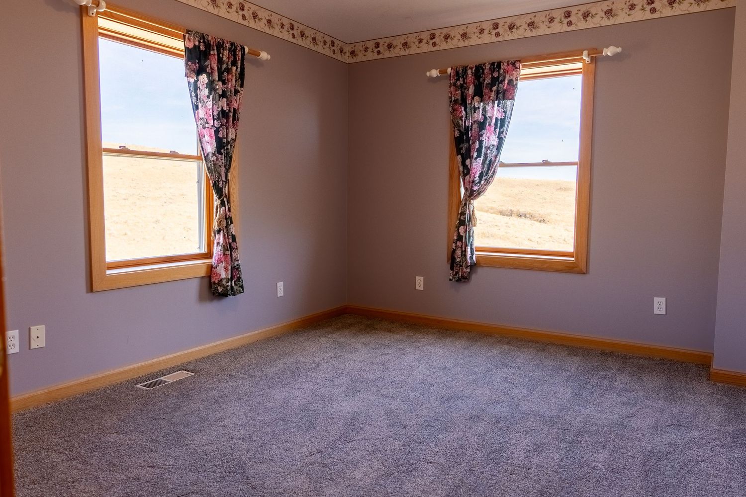 Empty bedroom with gray carpet, floral curtains, two windows, and beige walls with wood trim.