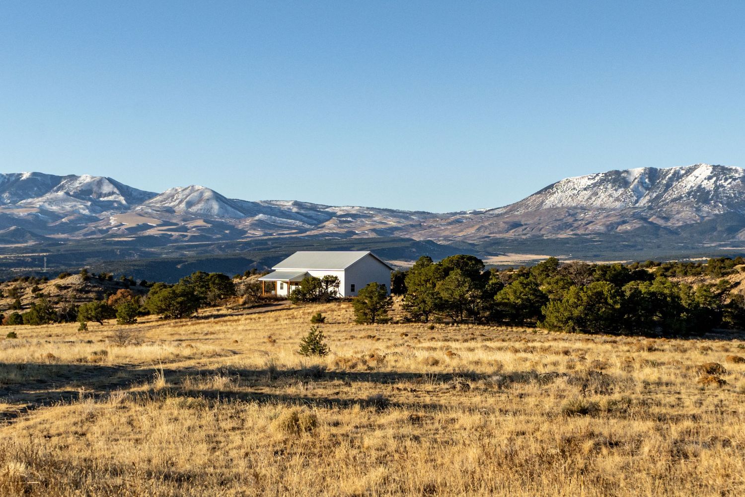 A white ranch house sits amid golden grasslands with snow-capped mountains and clear blue skies in the background.