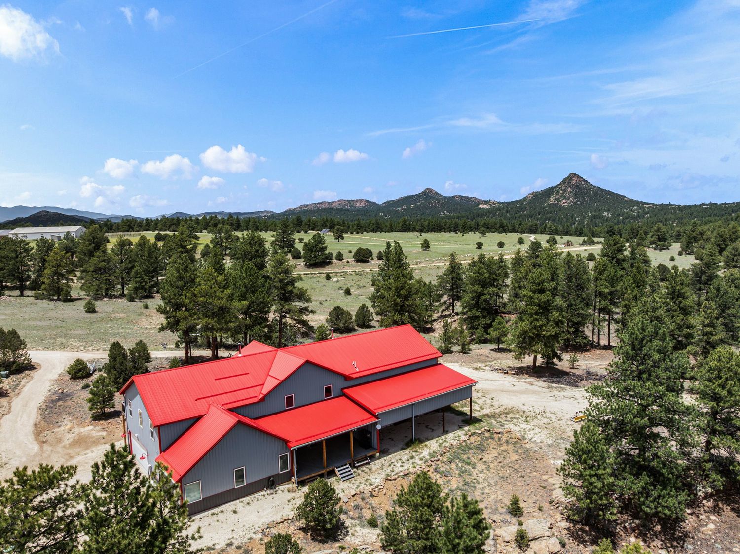Aerial view of red-roofed metal barn home surrounded by pine trees with dramatic mountain peaks in background.