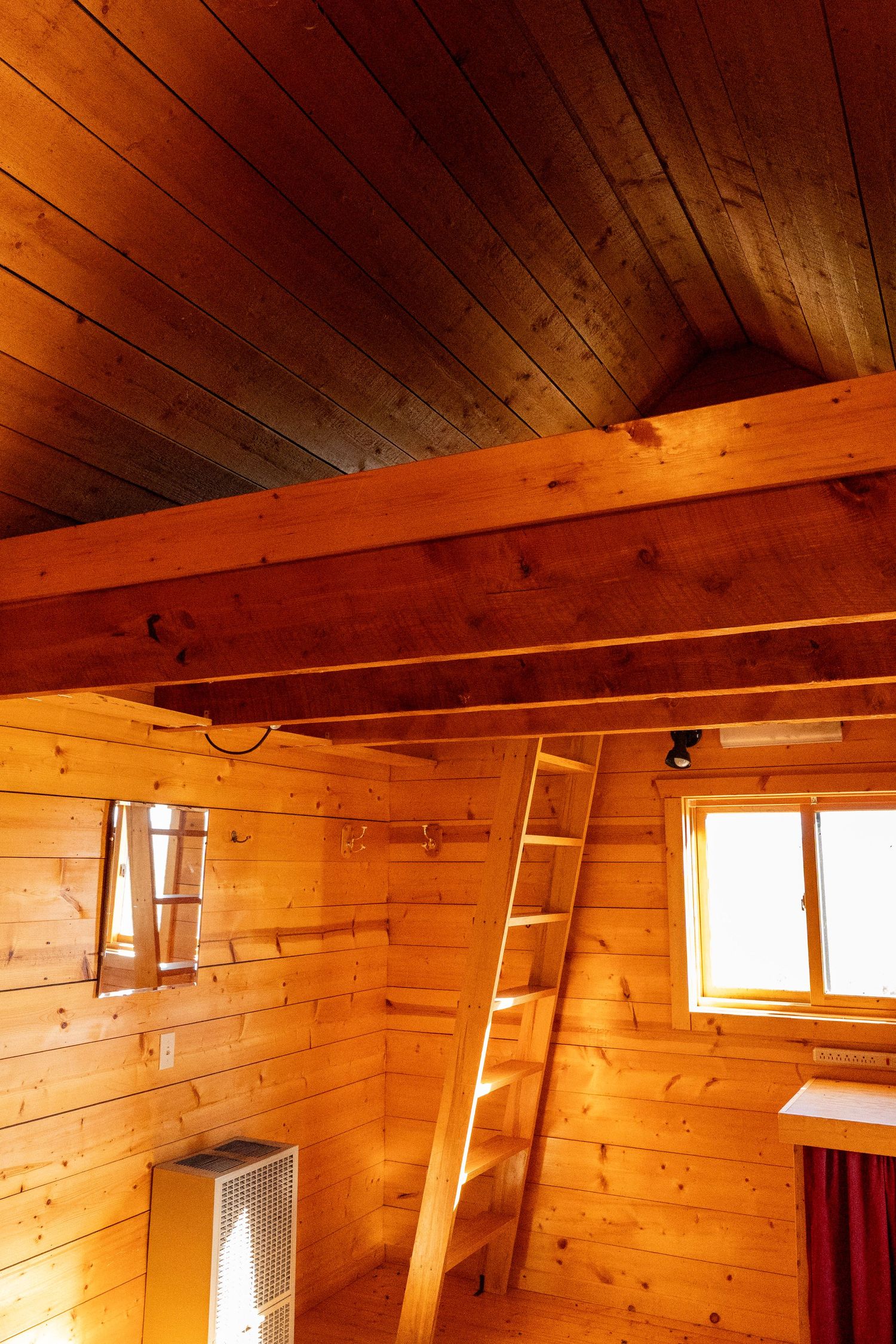 Rustic wooden cabin interior with exposed beam ceiling, ladder to loft, and warm amber tones throughout walls and ceiling.