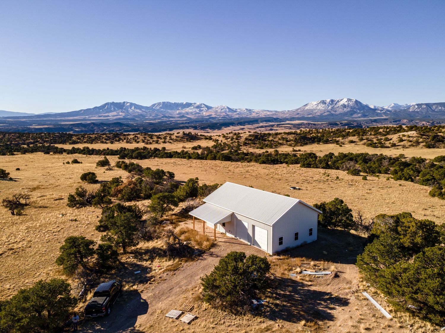 Aerial view of a white barn on golden plains with snow-capped mountains and scattered juniper trees under a clear blue sky.