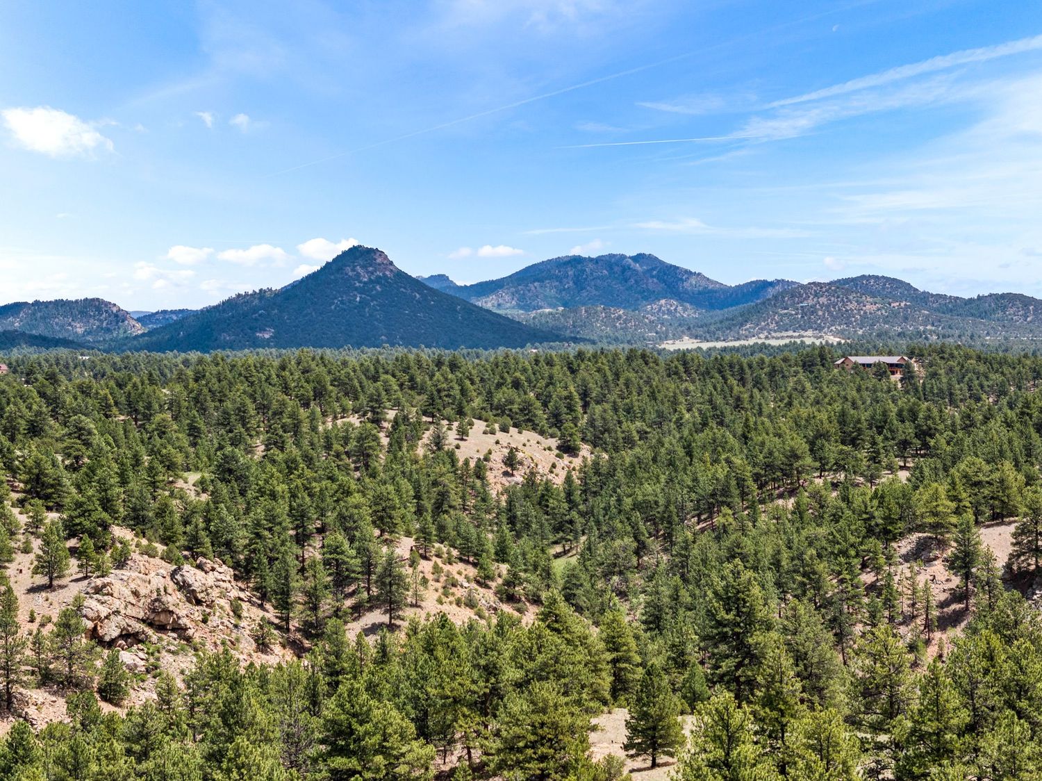Aerial view of dense pine forest with rocky outcrops and blue mountain peaks under a partly cloudy sky in Colorado.