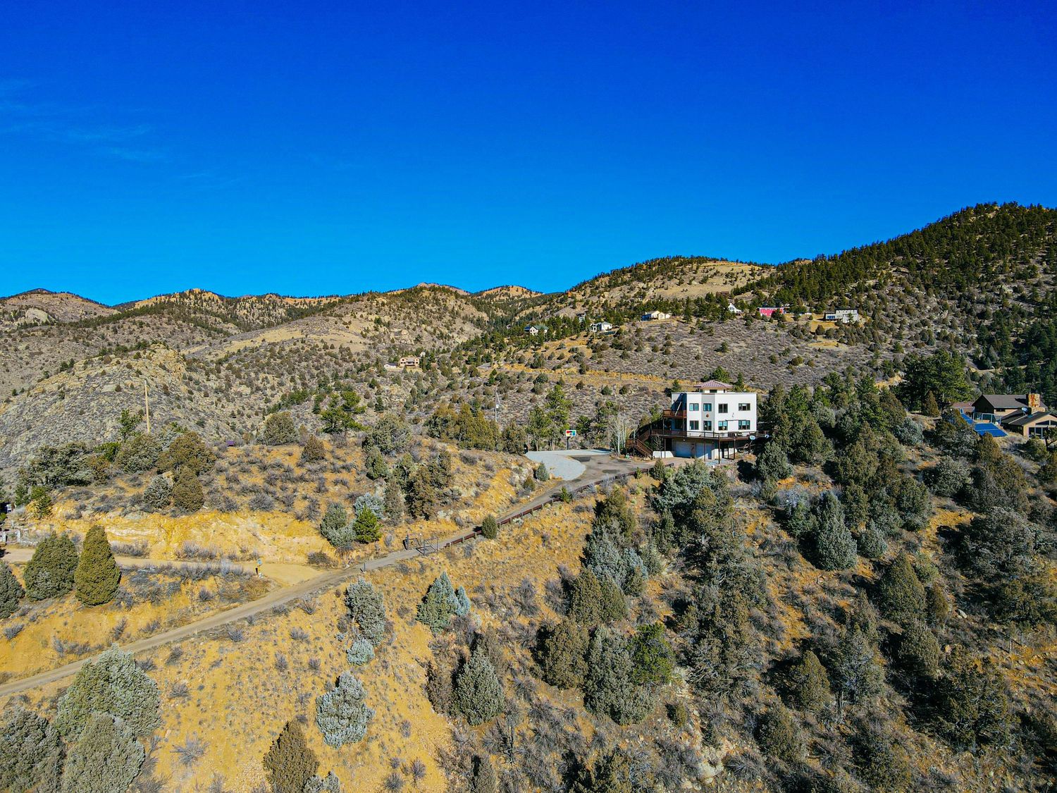 Aerial view of a white house nestled among evergreen trees on a rocky mountain hillside under a vivid blue sky.