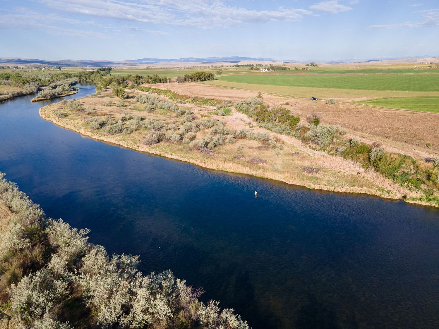 Aerial view of a calm river bend bordered by trees and green farmland under a clear blue sky.
