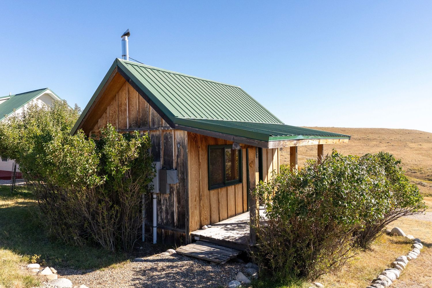 Rustic wooden cabin with green metal roof surrounded by shrubs in an open grassy landscape under clear blue sky.