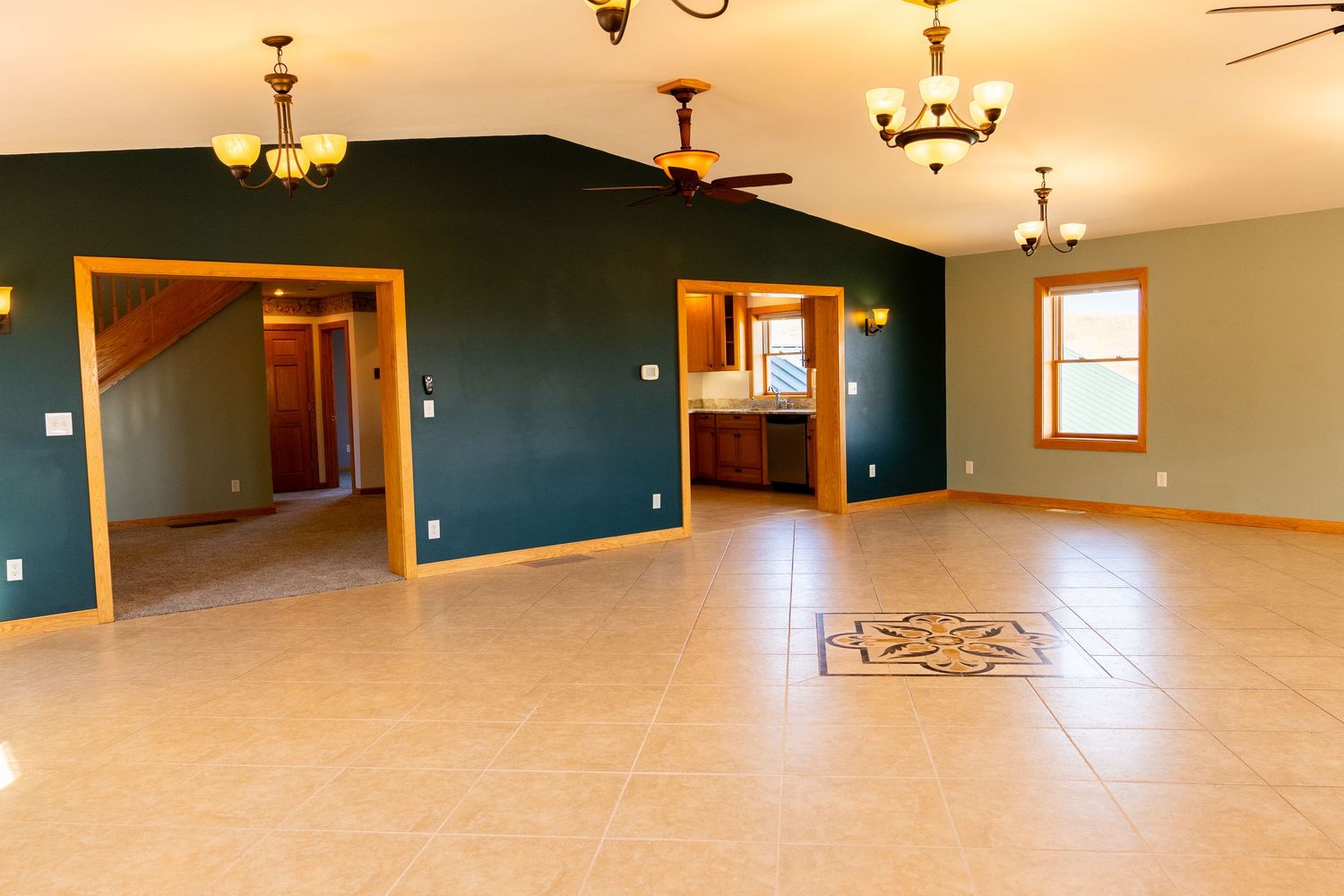 Spacious open-concept living room with teal walls, tile flooring, vaulted ceilings, and multiple chandeliers in a modern home.