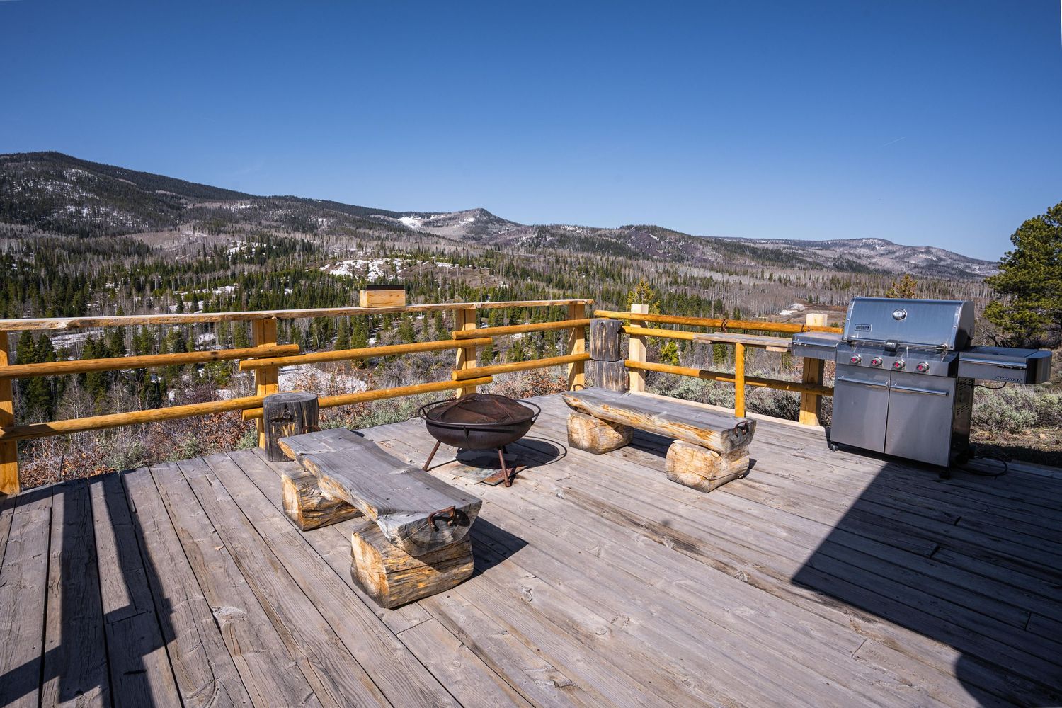 Spacious wooden deck with fire pit, benches, and grill overlooking a stunning mountain and forest  on the Colorado landscape.
