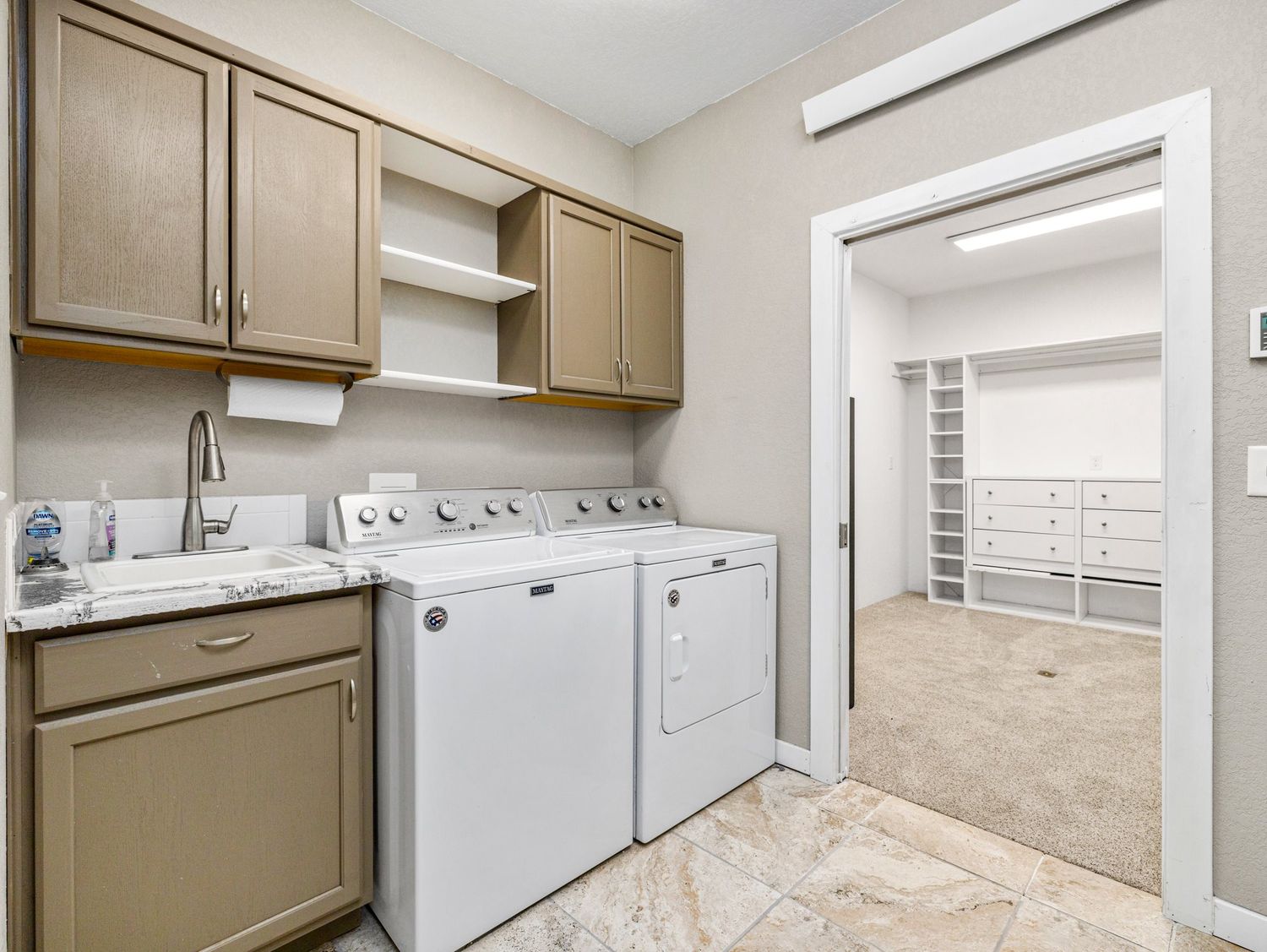 Spacious laundry room with white washer and dryer, wood cabinets, utility sink, and view into walk-in closet.