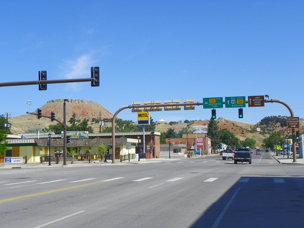 A wide main street intersection with traffic signals and highway signs in a small western town with hills in the background.