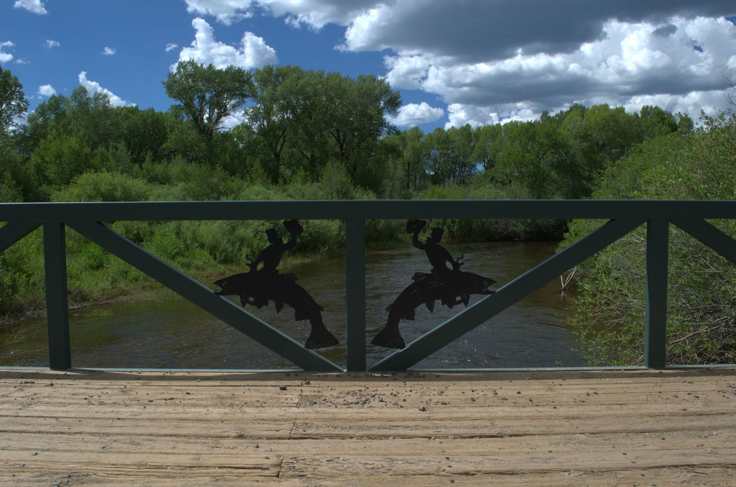Bridge railing with decorative metal fish cutouts overlooking a scenic river surrounded by lush green trees on luxury Wyoming property.