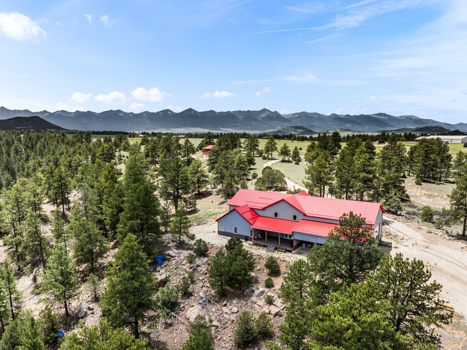 Aerial view of a red-roofed ranch house surrounded by pine trees with scenic mountain range backdrop under blue skies.