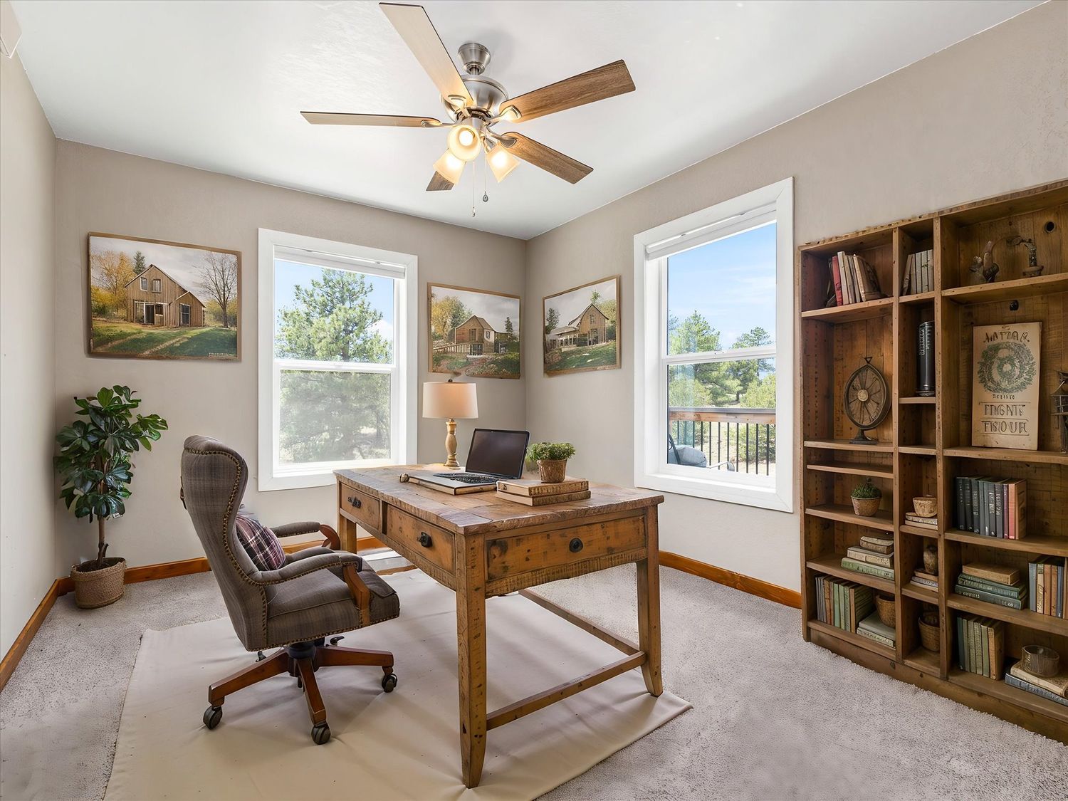 Cozy home office with wooden desk, bookshelf, ceiling fan, and natural light from two windows on beige carpet.