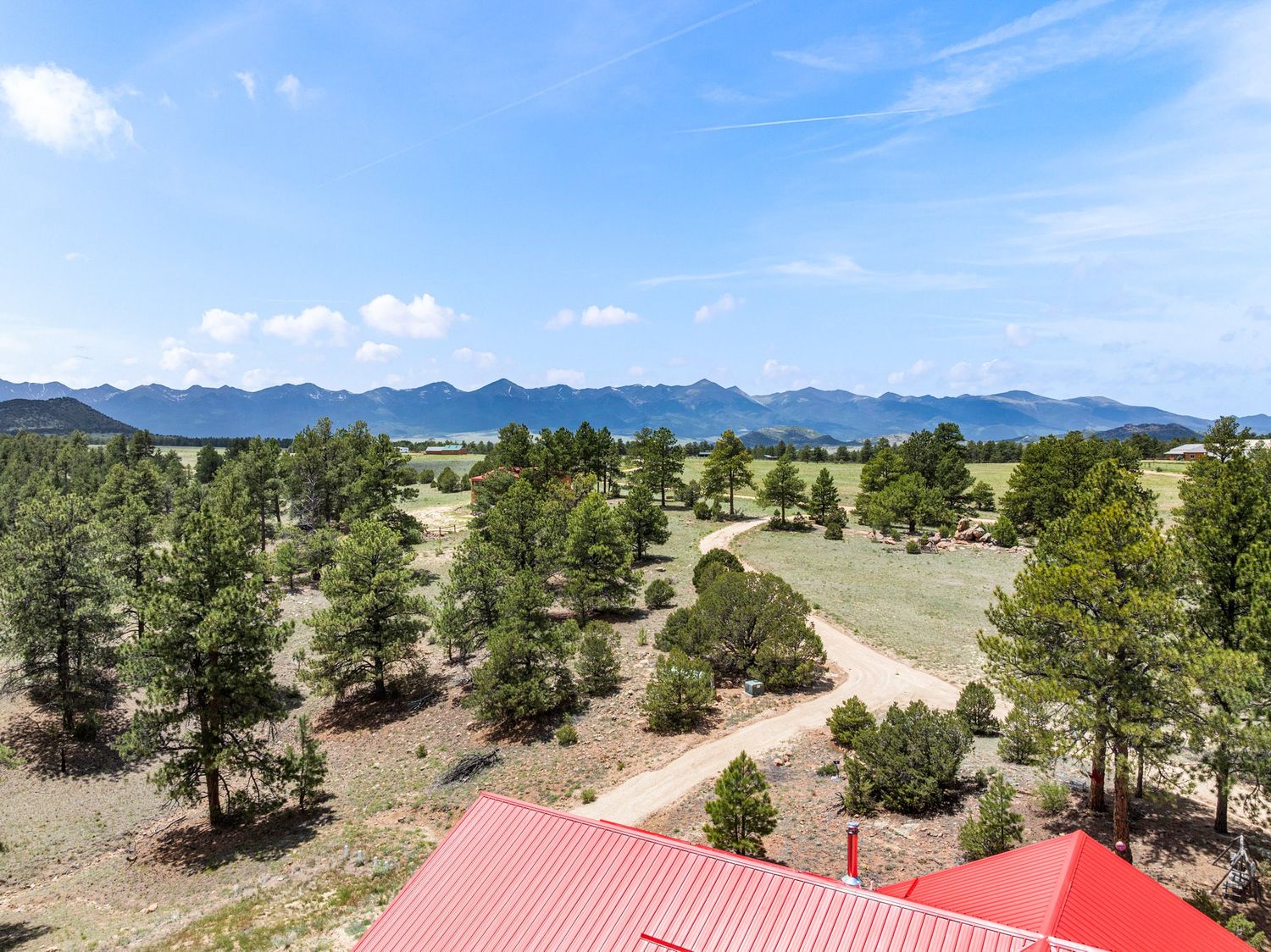 Aerial view of a rural property with pine trees, dirt road, red roof, and stunning mountain range backdrop under blue skies.