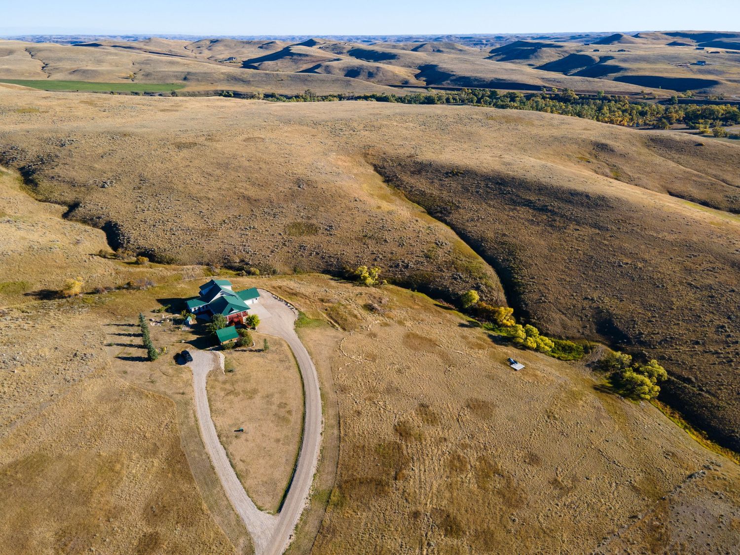 Aerial view of a remote farmhouse with a winding dirt driveway surrounded by vast rolling golden prairies under a clear sky.