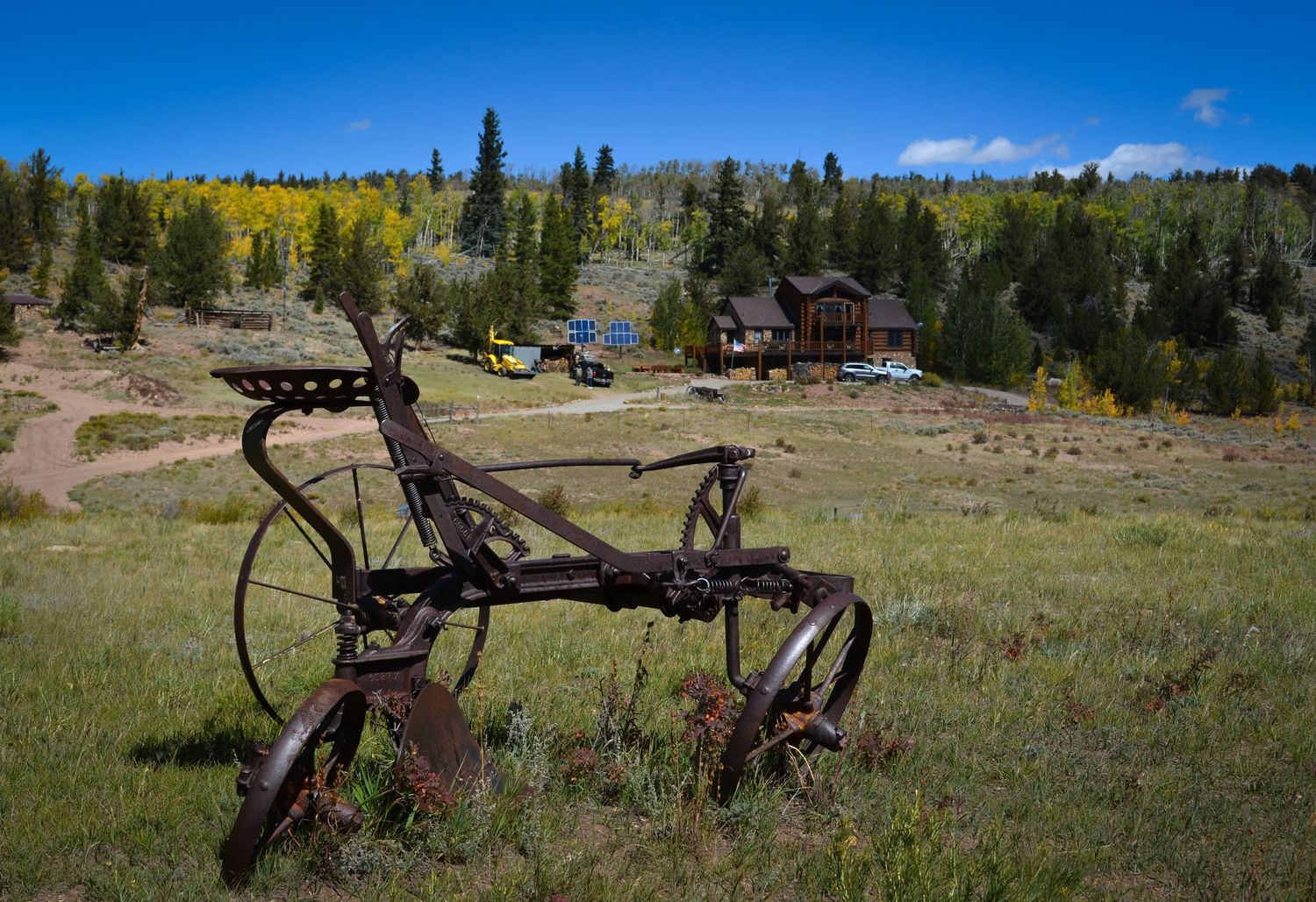 Antique farm plow in a mountain meadow with autumn trees and a luxury wooden cabin in the background under blue skies.