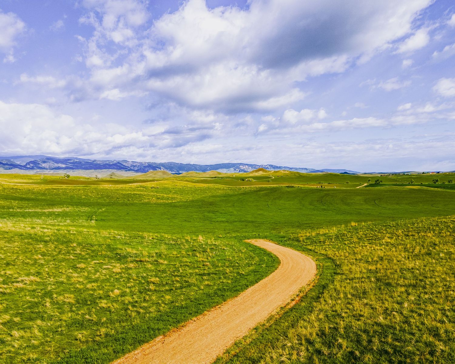 Winding dirt path through rolling green hills and grasslands under cloudy blue skies.