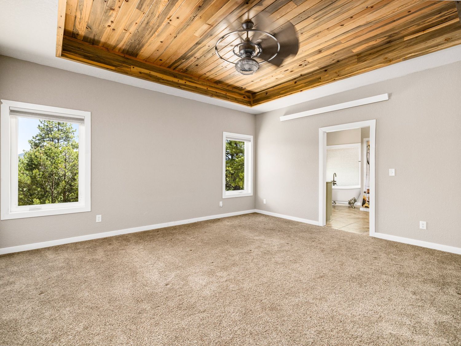 Spacious bedroom with plush carpet, wooden tray ceiling, ceiling fan, and natural light from windows.