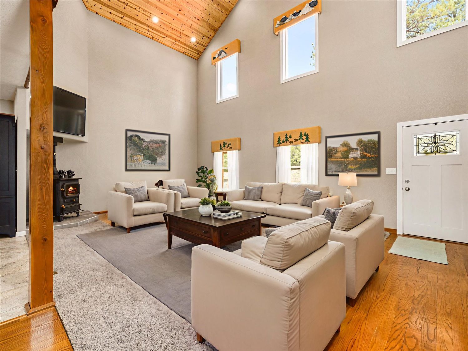 Spacious living room with vaulted wood ceiling, large windows, neutral sofas, hardwood floors, and a wood-burning stove.