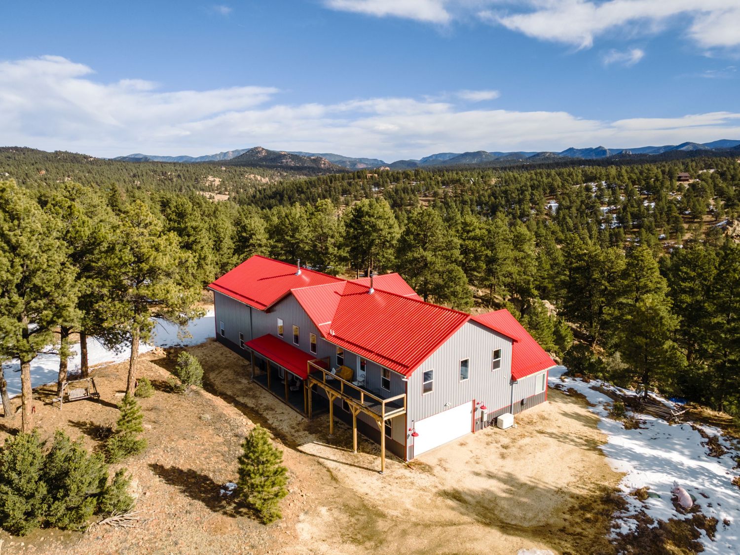 Aerial view of mountain home with red roof surrounded by pine forest and patches of snow.
