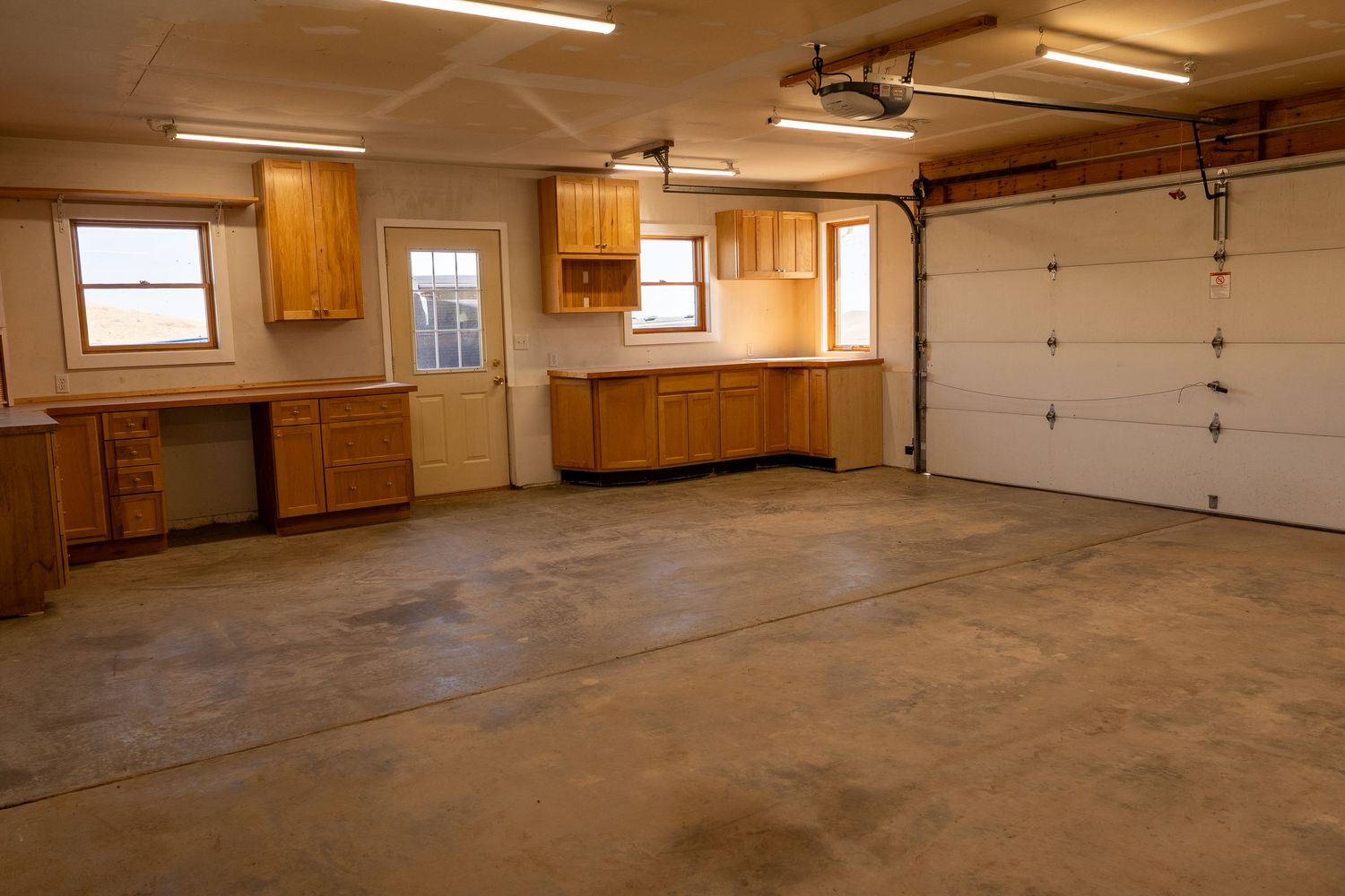 Spacious garage workshop with wooden cabinets, built-in desk, concrete floor, and natural light from two windows.