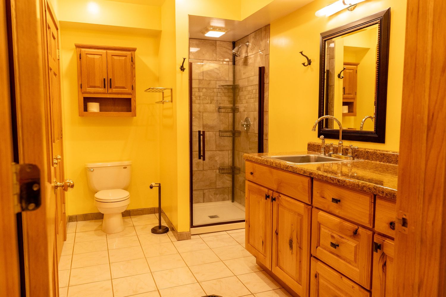 Spacious bathroom with wood cabinets, granite countertop, glass shower, white tile floors, and warm yellow walls.