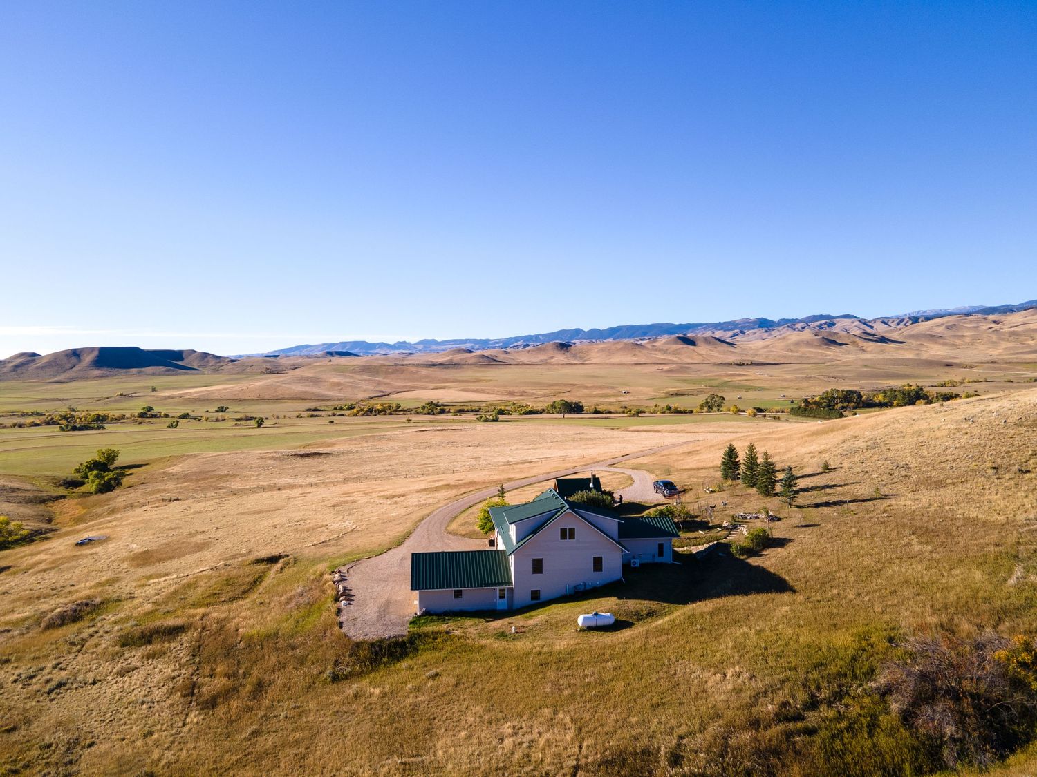 Aerial view of an isolated farmhouse surrounded by vast golden grasslands with rolling mountains under a clear blue sky.