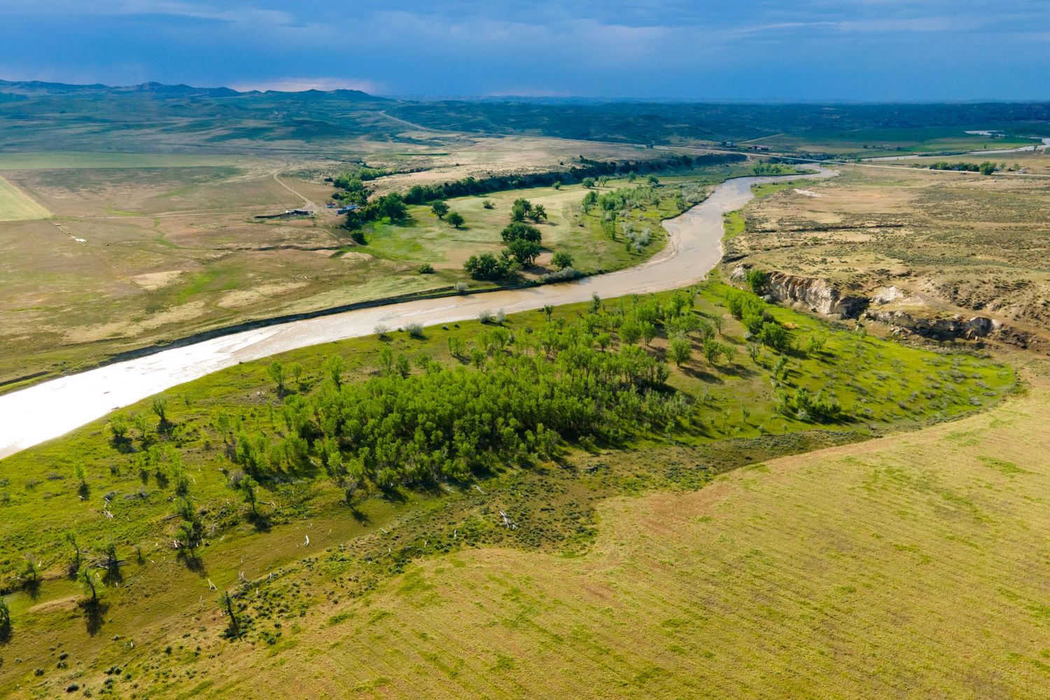 Aerial view of a winding river through vast green ranch land and rugged terrain under a cloudy sky in Wyoming.