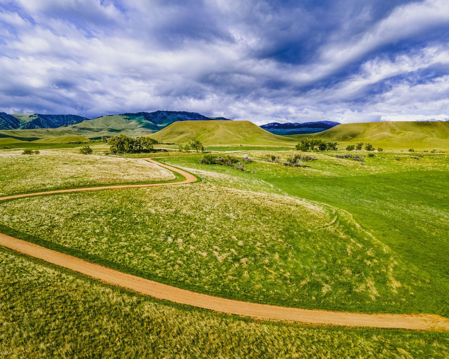 Drone shot of a winding dirt road cutting through green prairie grasslands beneath dramatic skies.