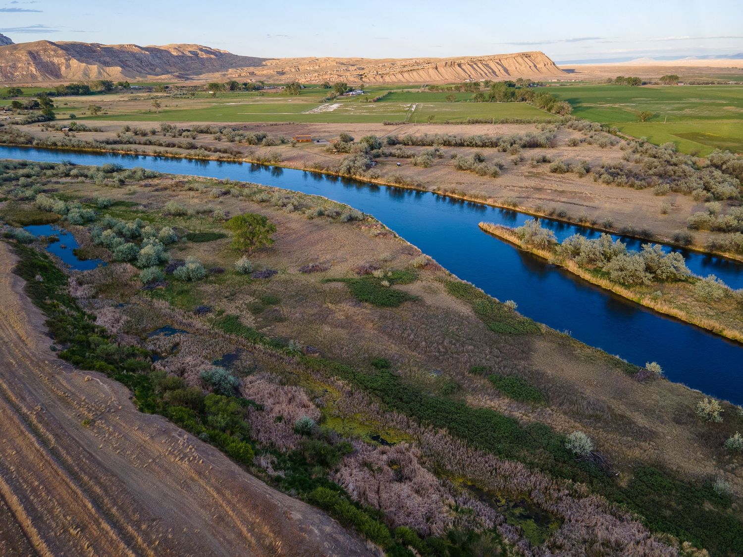 Aerial view of a blue irrigation canal winding through green farmland beside red cliffs at golden hour.