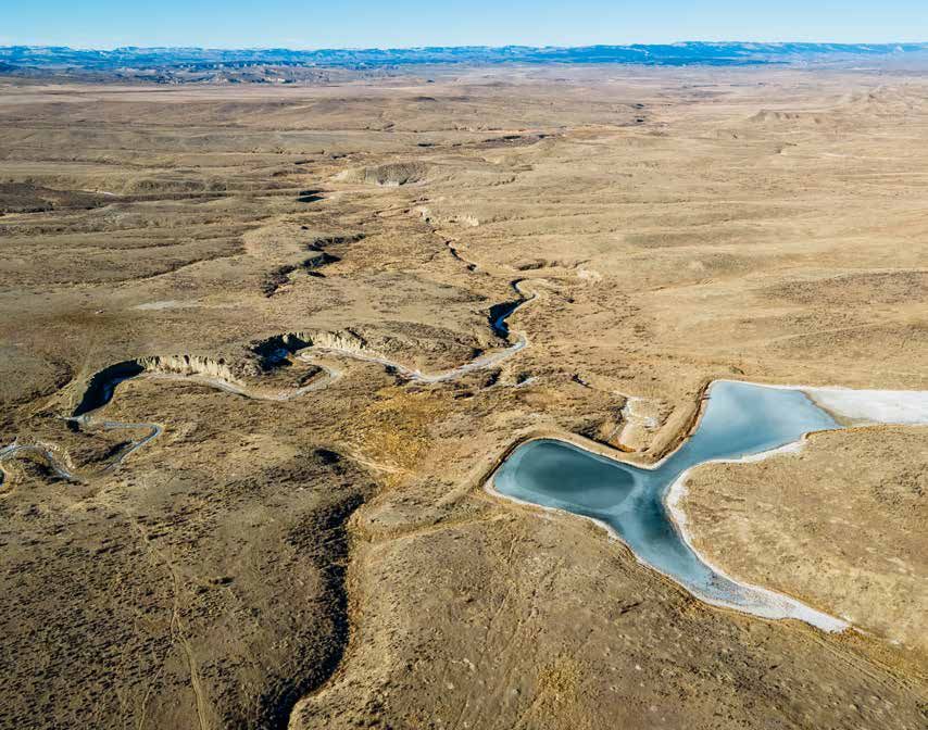 Aerial view of arid desert landscape with a winding canyon, small reservoir, and vast plains under a clear sky.