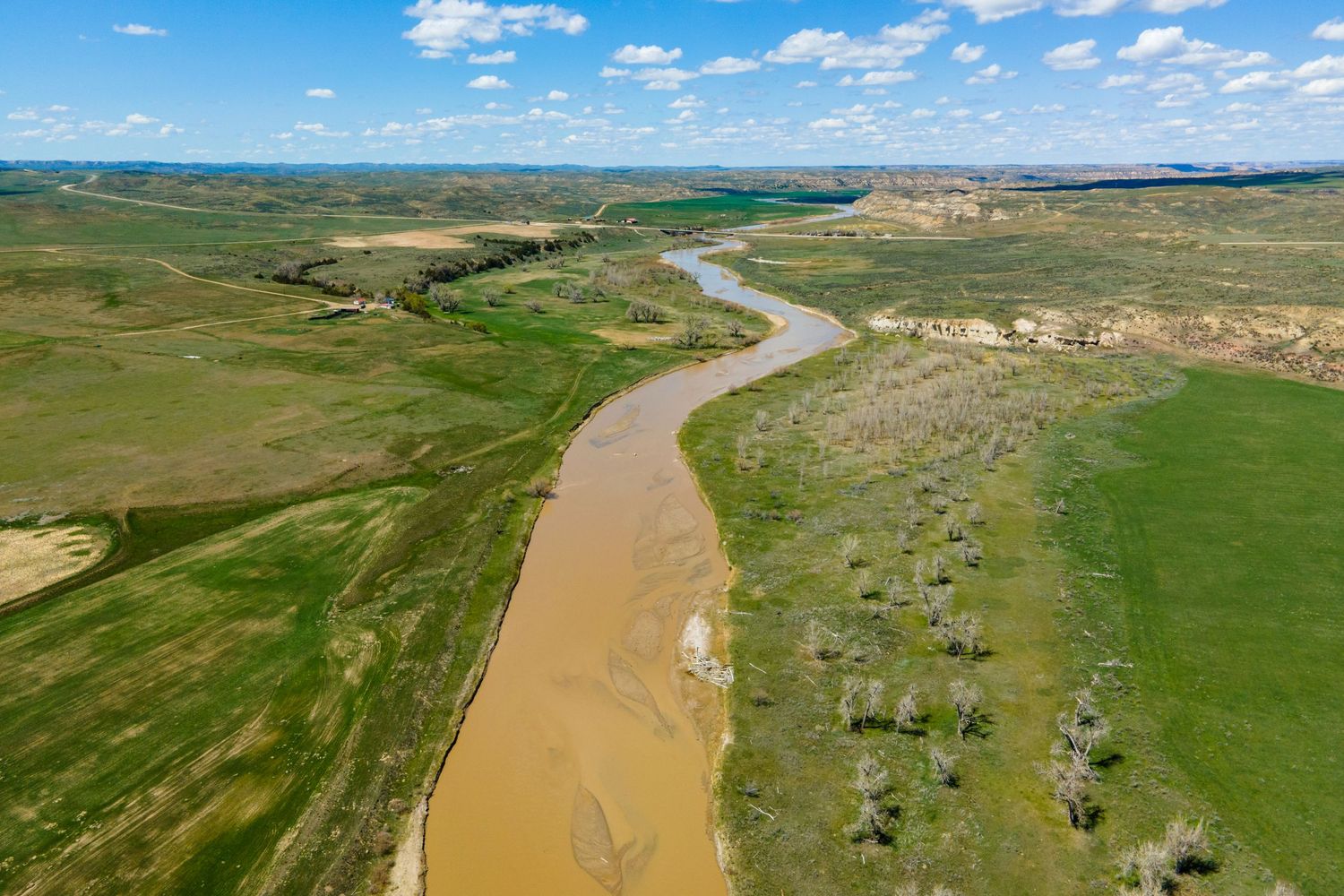 Aerial view of a winding Wyoming river through green rolling plains under a partly cloudy blue sky.