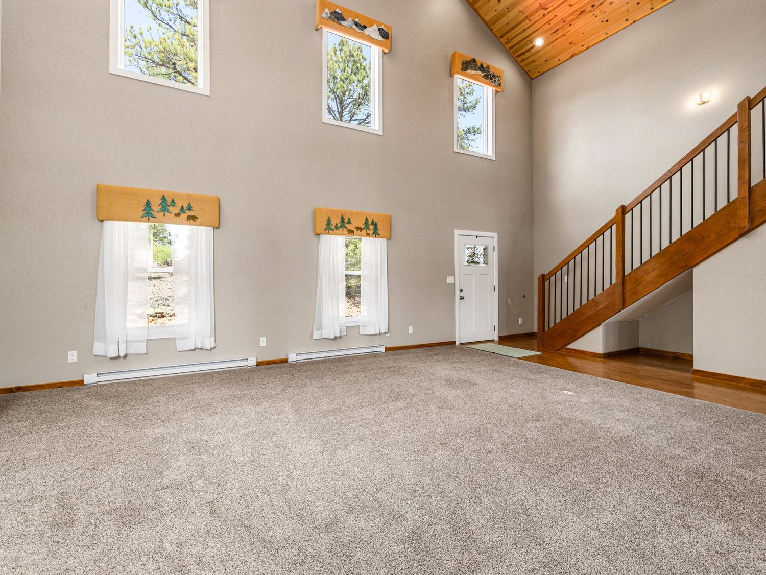 Spacious living room with vaulted wood ceiling, large windows, gray carpet, and open staircase with wood railing.