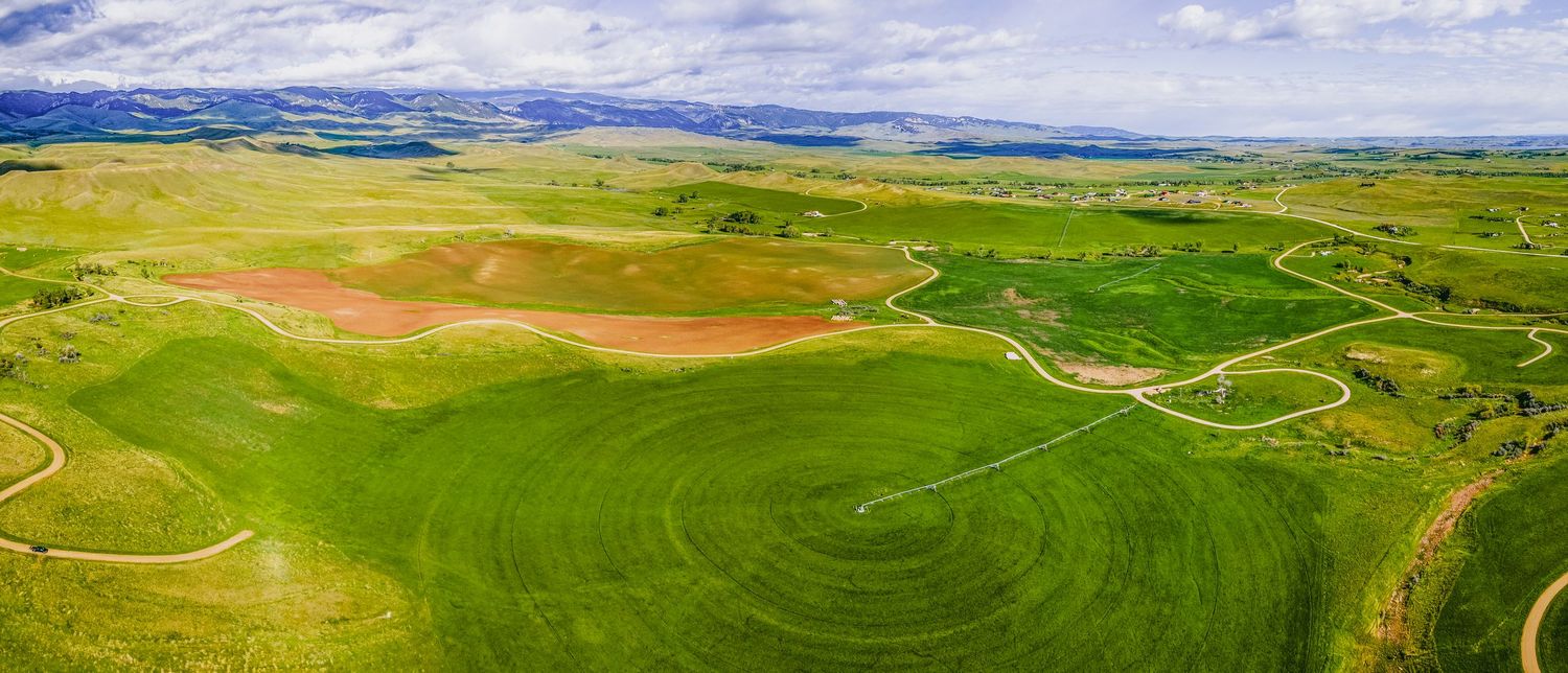 Wide panorama of lush farmland with meandering trails and mountains under a cloudy blue sky.