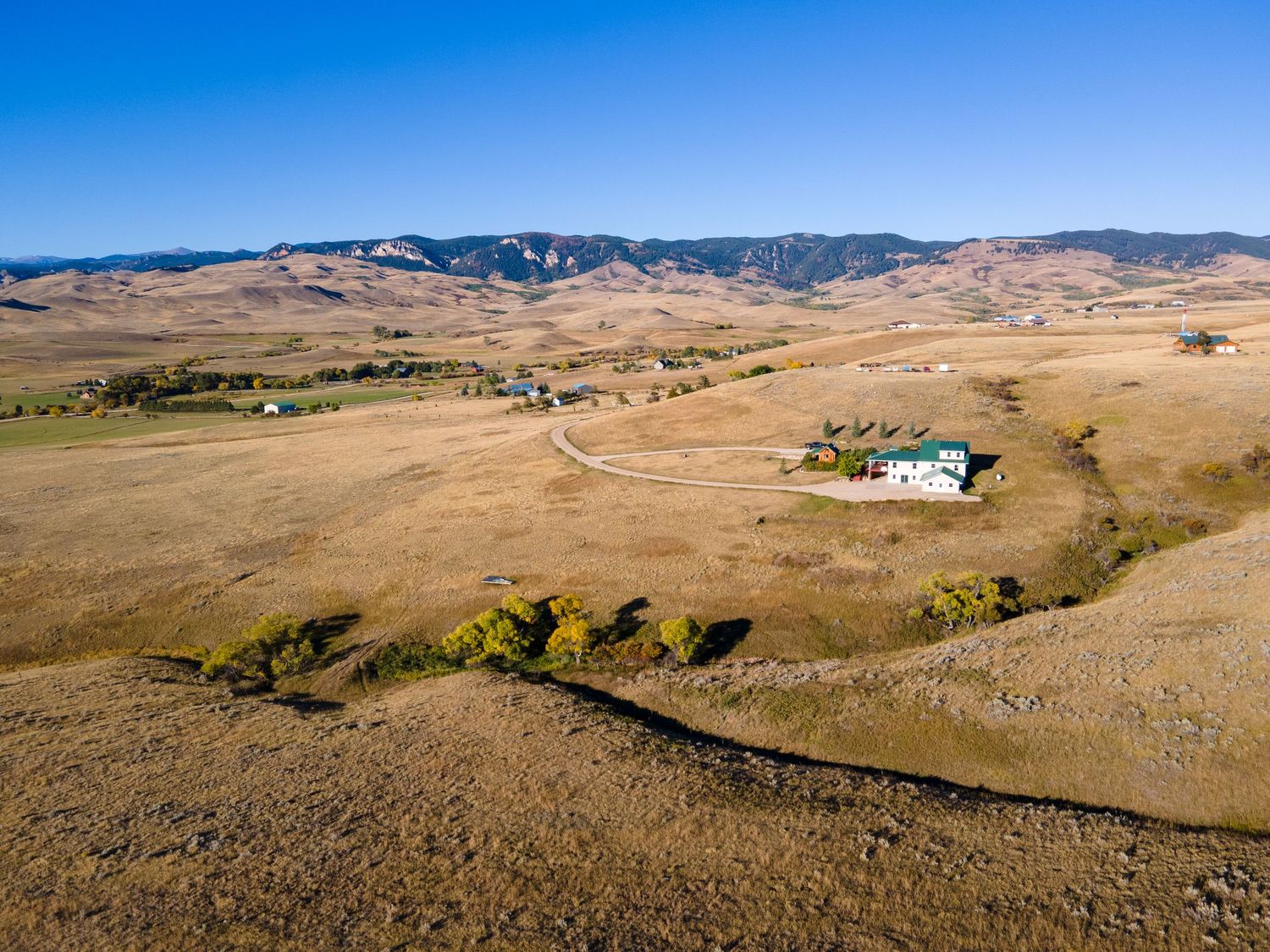 Aerial view of a rural ranch with a white farmhouse surrounded by golden grasslands and rolling mountains under a clear blue sky.