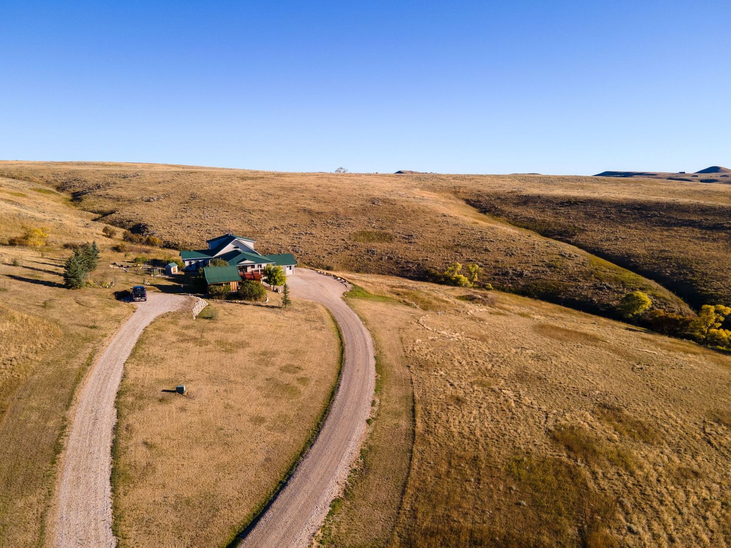 Aerial view of a rural homestead with winding dirt road through golden grasslands under a clear blue sky.