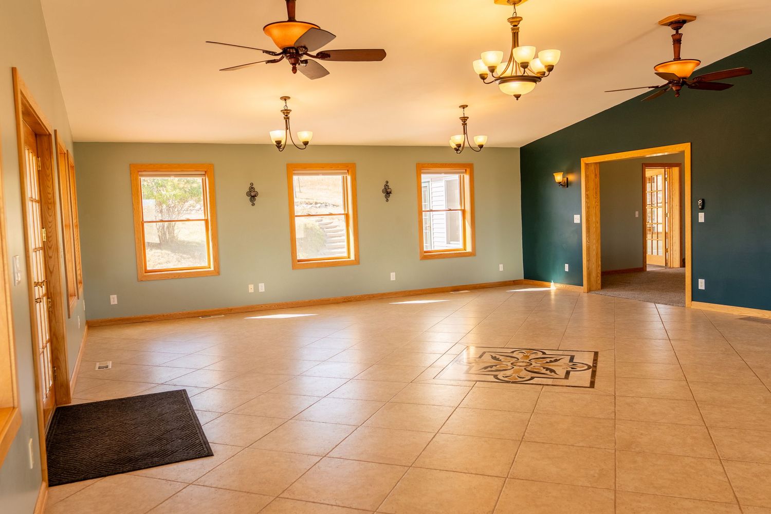 Spacious living room with teal walls, tile floors, ceiling fans, and large windows providing natural light.