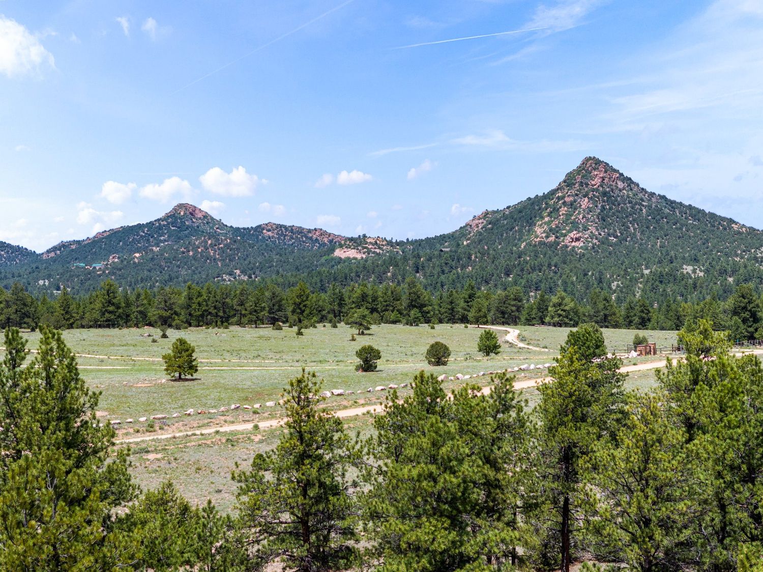 Scenic Colorado mountain landscape with twin rocky peaks, lush pine forest, and open green meadow under a blue sky.