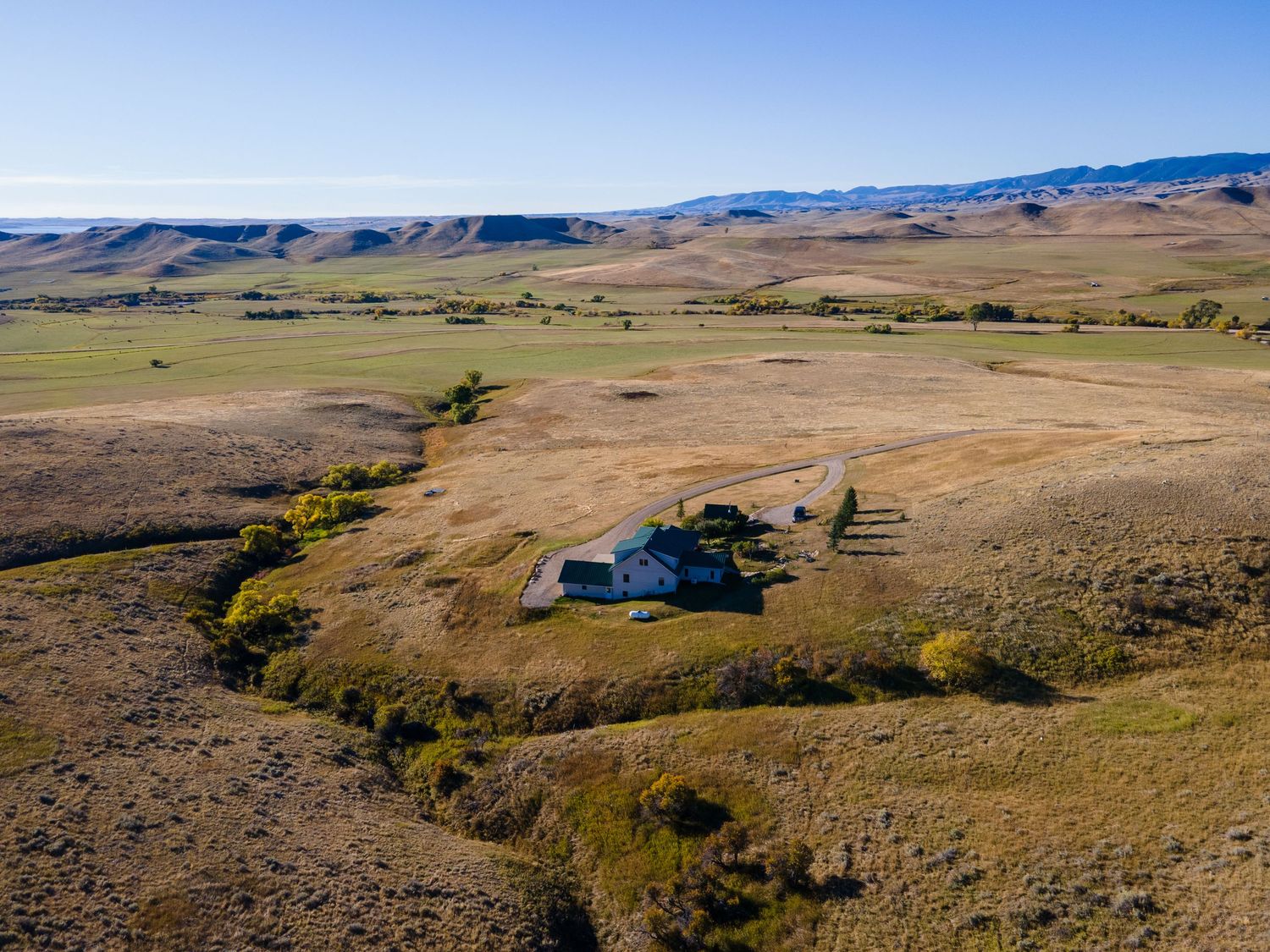 Aerial view of a remote farmstead surrounded by vast golden grasslands and rolling hills under a clear blue sky.