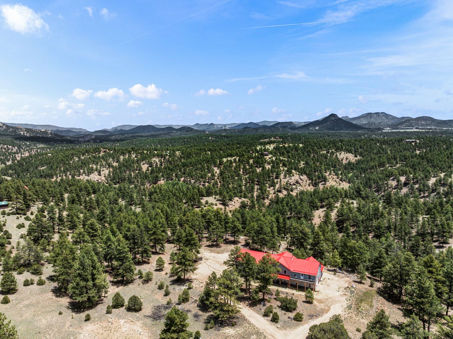 Aerial view of a secluded mountain home with red roof surrounded by dense pine forest and rocky peaks under blue sky.