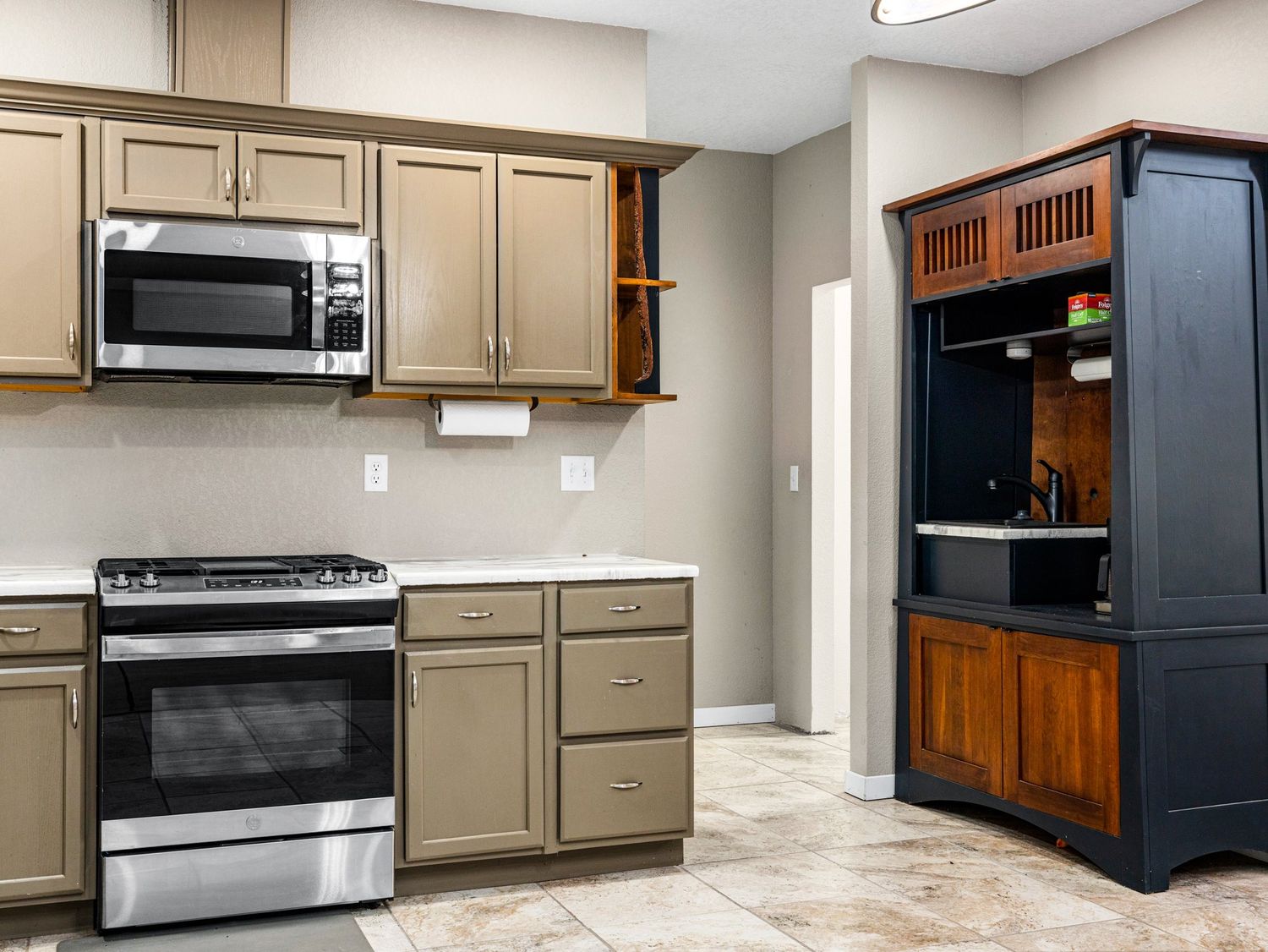 Modern kitchen with stainless steel gas range, microwave, taupe cabinets, and a dark wood entertainment armoire.