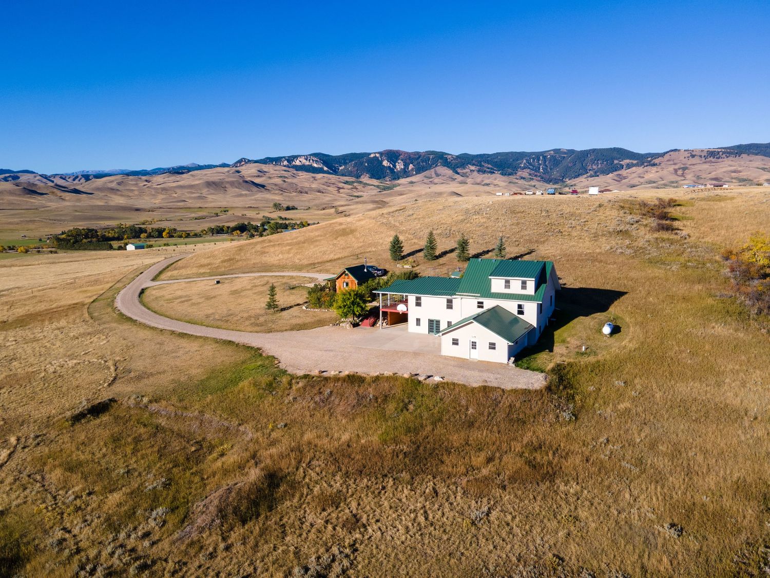 Aerial view of a rural farmhouse with green roof surrounded by golden fields and mountain backdrop under clear blue skies.
