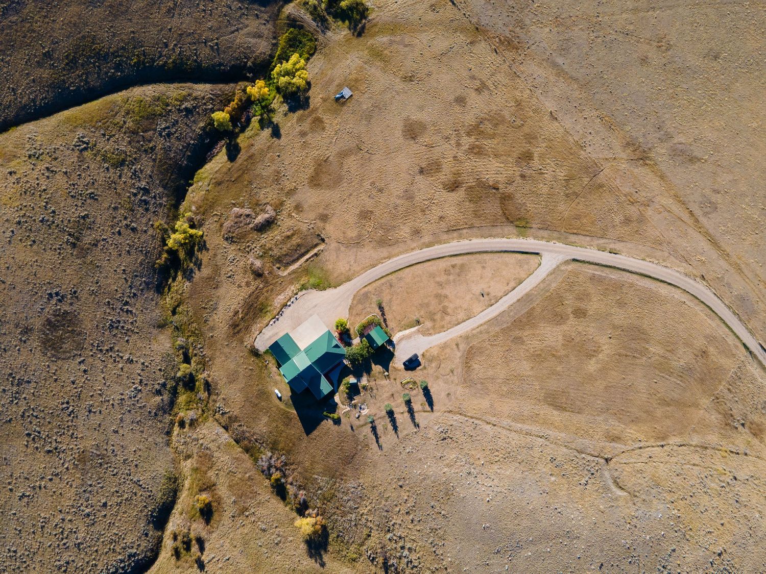 Aerial drone view of a rural homestead with green-roofed buildings on arid brown landscape with sparse trees.