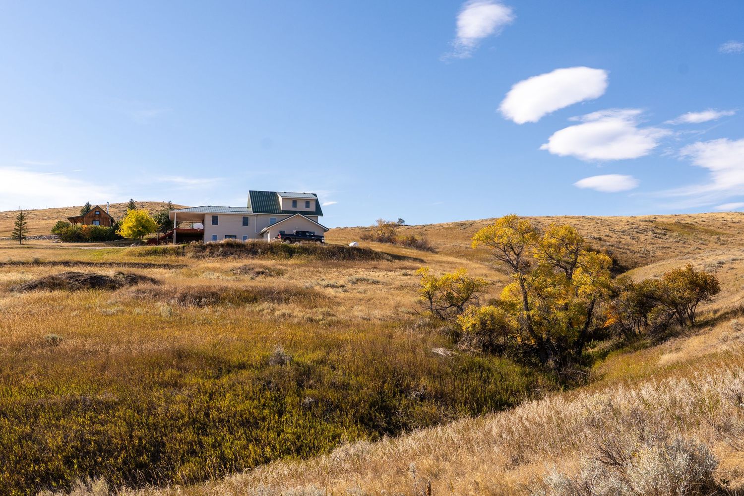 A rustic farmhouse with a green roof sits among golden autumn grasslands under a bright blue sky in a rural prairie landscape.