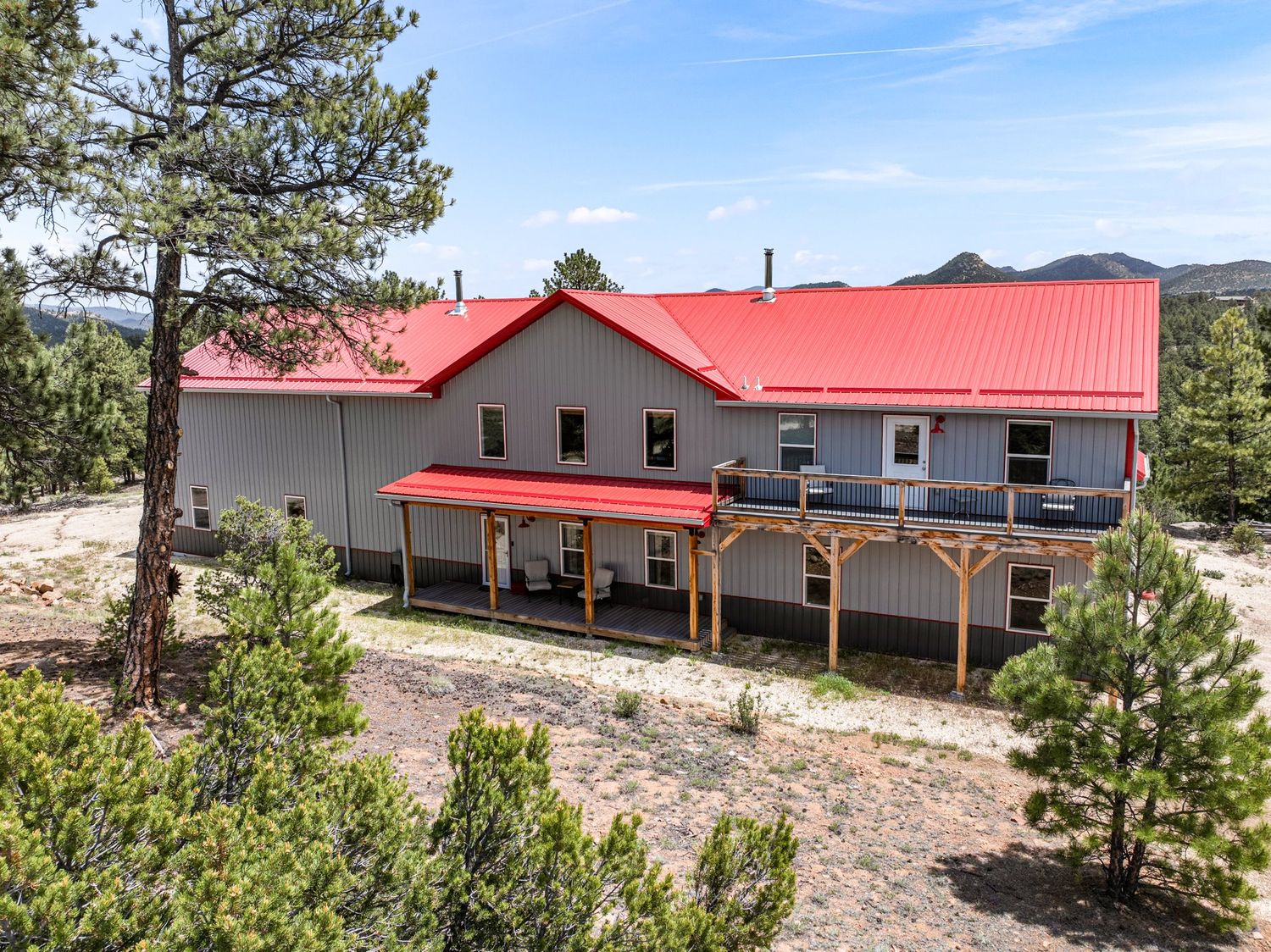 Exterior of two-story home with red metal roof, covered porch, wood deck, and pine tree surroundings.