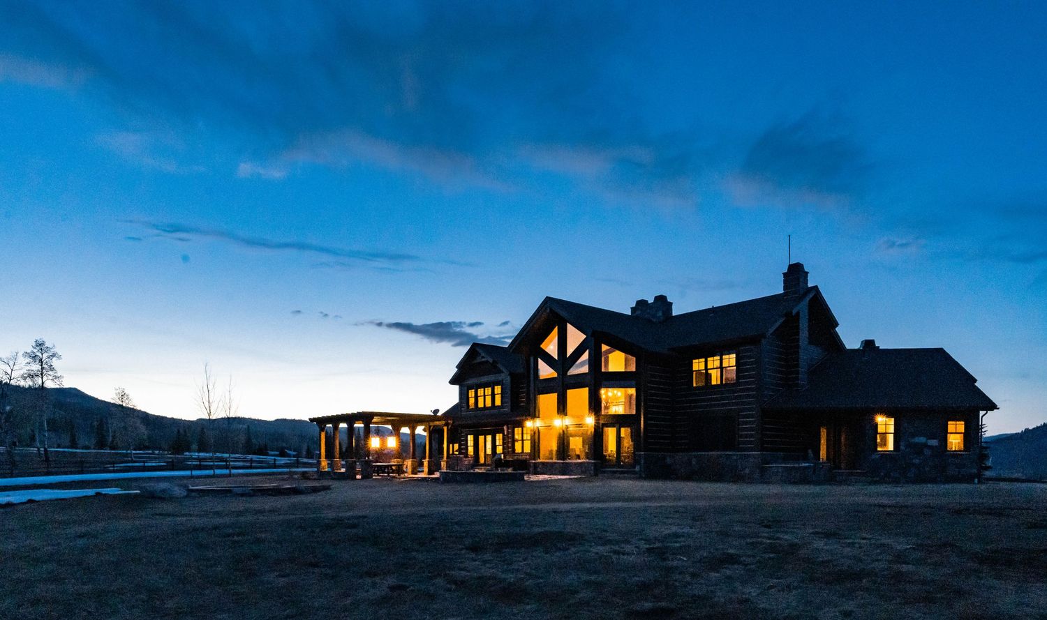 Large log cabin home illuminated at dusk with warm glowing windows against a twilight mountain sky in Colorado.